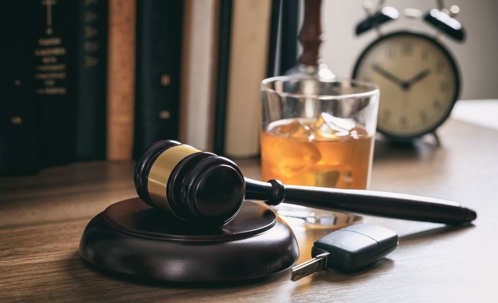 A glass of an alcoholic beverage sitting between legal books, an alarm clock, a judge's gavel, and a car key.